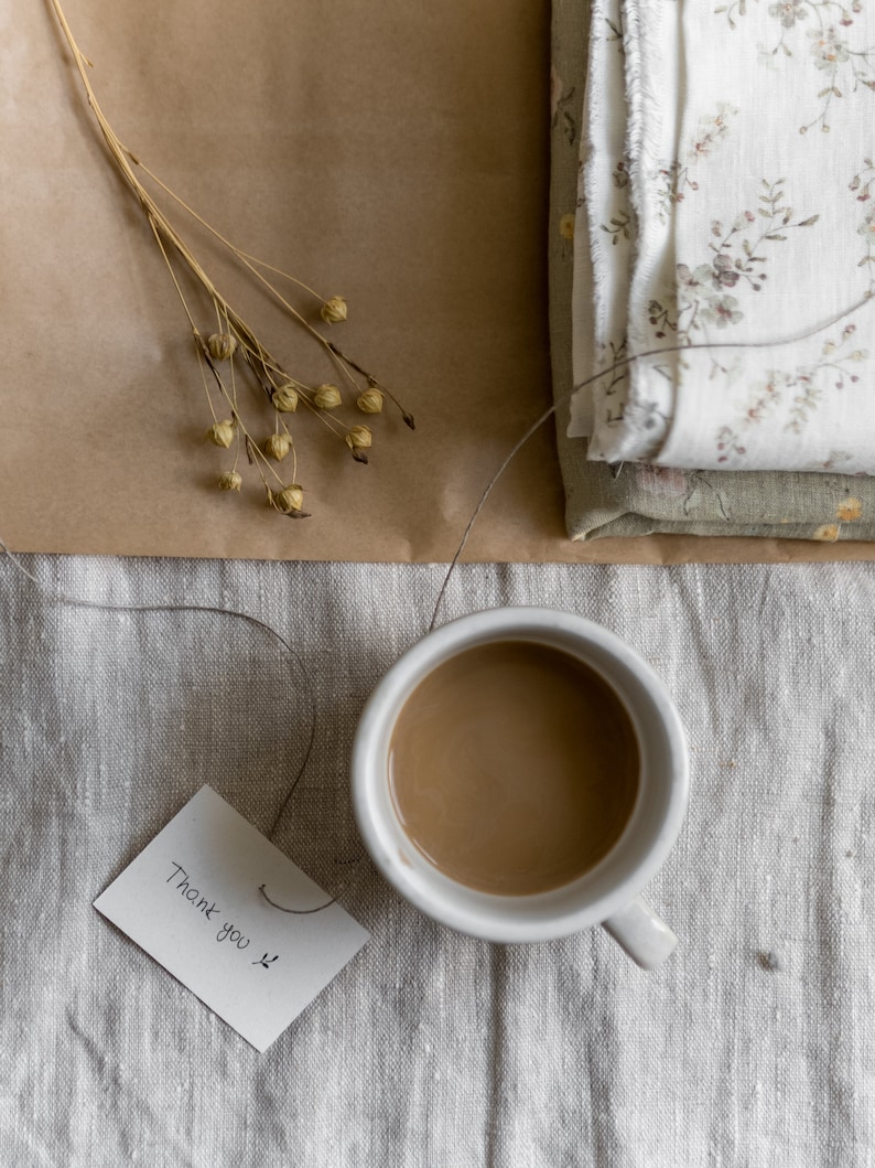Puede incluir: Una taza de cer&aacute;mica blanca llena de caf&eacute; se encuentra sobre un mantel de lino beige. Una peque&ntilde;a tarjeta blanca con el texto "Gracias" est&aacute; sobre la mesa junto a la taza.