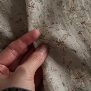 Könnte beinhalten: Ein hellsalbeigrüner Stoff mit einem sich wiederholenden Blumenmuster in sanften Beige- und gedeckten Brauntönen. Das Textil weist eine sichtbare Webstruktur mit matter Oberfläche auf und zeigt zarte Blumenillustrationen.