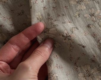 May include: A light sage green fabric featuring a repeating floral pattern in soft beige and muted brown tones. The textile has a visible woven texture with a matte finish, showcasing delicate flower illustrations across the entire surface.