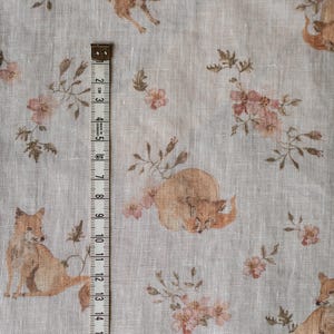 Puede incluir: Una tela de lino beige con un patr&oacute;n repetido de zorros y flores rosas. Una cinta m&eacute;trica se coloca verticalmente en el lado izquierdo de la tela.