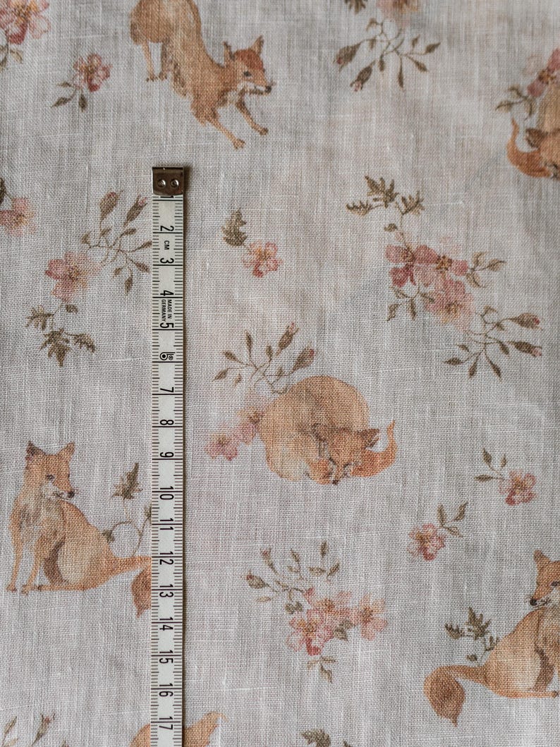 Puede incluir: Una tela de lino beige con un patr&oacute;n repetido de zorros y flores rosas. Una cinta m&eacute;trica se coloca verticalmente en el lado izquierdo de la tela.