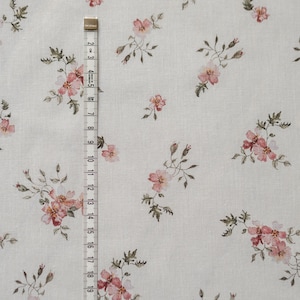 K&ouml;nnte beinhalten: Ein wei&szlig;er Stoff mit einem zarten Blumenmuster mit kleinen rosa Bl&uuml;ten und gr&uuml;nen Bl&auml;ttern. Der Stoff wird mit einem Ma&szlig;band gemessen.
