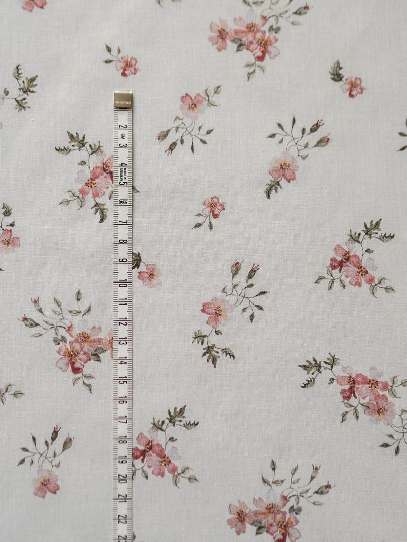 K&ouml;nnte beinhalten: Ein wei&szlig;er Stoff mit einem zarten Blumenmuster mit kleinen rosa Bl&uuml;ten und gr&uuml;nen Bl&auml;ttern. Der Stoff wird mit einem Ma&szlig;band gemessen.