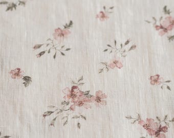 May include: A light beige fabric featuring a repeating pattern of small, delicate pink flowers with muted green leaves and stems. The textile has a visible woven texture with a subtle, natural grain throughout the surface of the material.