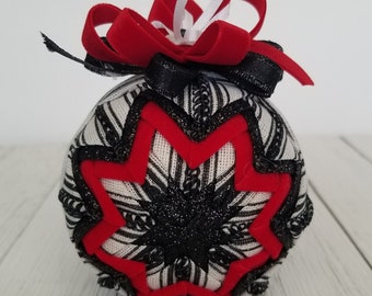 May include: A decorative Christmas ornament with a black and white patterned fabric base. The ornament features a red starburst design and is accented with black glitter and red velvet ribbon. A white ribbon loop is attached for hanging.
