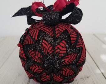 May include: A red and black Christmas ornament with a black and red ribbon bow. The ornament is made of fabric and has a glittery finish.
