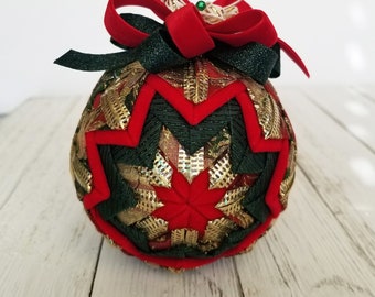 May include: A handcrafted Christmas ornament with a starburst design. The ornament features red, green, and gold fabrics, accented with a red velvet bow and a green bead. A white string loop is attached for hanging.