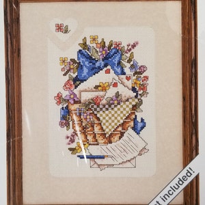 May include: A cross-stitch pattern featuring a basket overflowing with colorful flowers and a blue bow. The basket is filled with white envelopes and a pen. The text "mat included!" is in the lower right corner of the image.