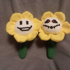 May include: Two plush yellow flower toys with white faces. One flower has a smiling face and the other has a frowning face with red eyes.