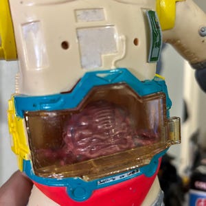 May include: Vintage action figure with a cream body, yellow and blue details, and red shorts. The figure features a clear compartment showcasing a detailed, reddish-brown brain. The toy is partially visible.