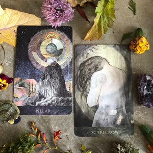 K&ouml;nnte beinhalten: Zwei Tarotkarten, eine mit dunkelblauem Hintergrund und einer gro&szlig;en grauen Felsformation mit dem Wort "PILLAR" unten, und die andere mit grauem Hintergrund und einer Figur, die dem Betrachter den R&uuml;cken zukehrt, mit dem Wort "SCARECROW" unten.