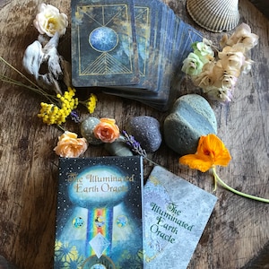 Może przedstawiać: Talia kart i przewodnik do wr&oacute;żb z  Illuminated Earth Oracle. Talia kart ma niebieski i złoty wz&oacute;r z motywem niebieskim. Przewodnik jest biały z zielonym tekstem. Talia kart i przewodnik są otoczone naturalnymi elementami, takimi jak kwiaty, pi&oacute;ra i kamienie.