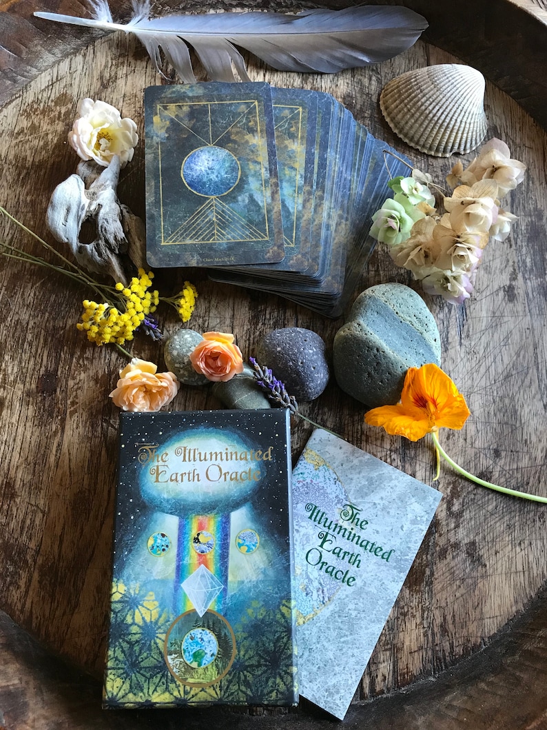 Może przedstawiać: Talia kart i przewodnik do wr&oacute;żb z  Illuminated Earth Oracle. Talia kart ma niebieski i złoty wz&oacute;r z motywem niebieskim. Przewodnik jest biały z zielonym tekstem. Talia kart i przewodnik są otoczone naturalnymi elementami, takimi jak kwiaty, pi&oacute;ra i kamienie.