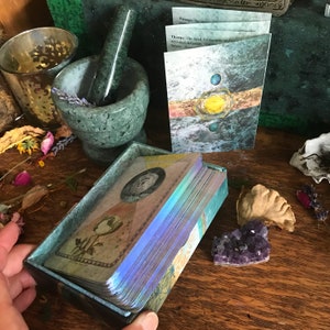 K&ouml;nnte beinhalten: Ein Tarotkarten-Deck in einer Schachtel mit einem blauen, gr&uuml;nen und violetten, schimmernden Irideszenz-Effekt. Die Schachtel ist ge&ouml;ffnet und die Karten sind aufgef&auml;chert. Die Karten haben eine Vintage-&Auml;sthetik mit Illustrationen von Blumen und Himmelsk&ouml;rpern. Der Text auf der Karte lautet "Dornen: Das Bed&uuml;rfnis nach Sicherheit und Grenzen, aktivierte Abwehrkr&auml;fte. Geh&auml;rtete Sch&ouml;nheit. Akzeptanz."