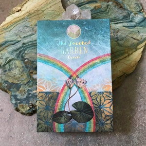 K&ouml;nnte beinhalten: Die Schachtel des Facettengarten-Orakel-Decks hat einen blaugr&uuml;nen Hintergrund mit einem doppelten Regenbogen und zwei wei&szlig;en Seerosen. Die Schachtel hat goldene Folienakzente und den Text "The Facetted Garden Oracle" in goldenen Buchstaben.