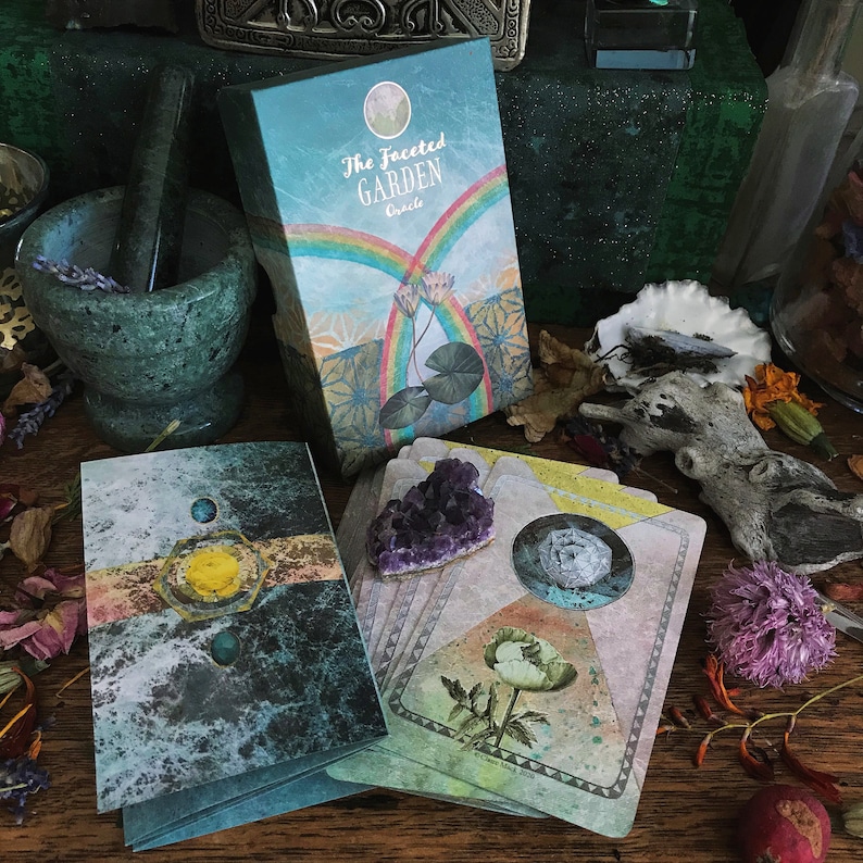 K&ouml;nnte beinhalten: Eine Schachtel mit The Faceted Garden Oracle-Karten, mit einem gr&uuml;n-gelb-blauen Design. Die Schachtel ist ge&ouml;ffnet und zeigt einen Stapel Karten mit einer wei&szlig;en Blume und einem Kristall darauf. Die Karten liegen auf einer Holzoberfl&auml;che mit anderen Dekorationsgegenst&auml;nden.