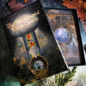 Może przedstawiać: Pudełko talii kart The Illuminated Earth Oracle ma ciemnoniebieskie tło z złotymi akcentami i wzorem tęczy. Pudełko jest zamknięte i widnieje na nim tytuł "The Illuminated Earth Oracle" złotymi literami.