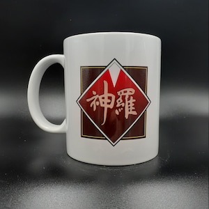 Könnte beinhalten: Weißer Keramikbecher mit großem Henkel. Der Becher hat eine braune und rote rautenförmige Grafik mit japanischen Schriftzeichen. Der Becher steht vor einem schwarzen Hintergrund.