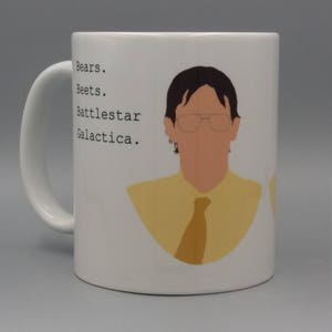 Puede incluir: Taza de cerámica blanca con una ilustración de dibujos animados de una persona con gafas y una camisa y corbata amarillas. La taza tiene las palabras "Bears. Beets. Battlestar Galactica."