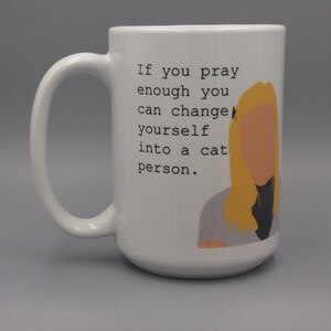 Puede incluir: Taza de cer&aacute;mica blanca con un asa grande. La taza presenta el texto "If you pray enough you can change yourself into a cat person." y una ilustraci&oacute;n de dibujos animados de una persona con cabello rubio.