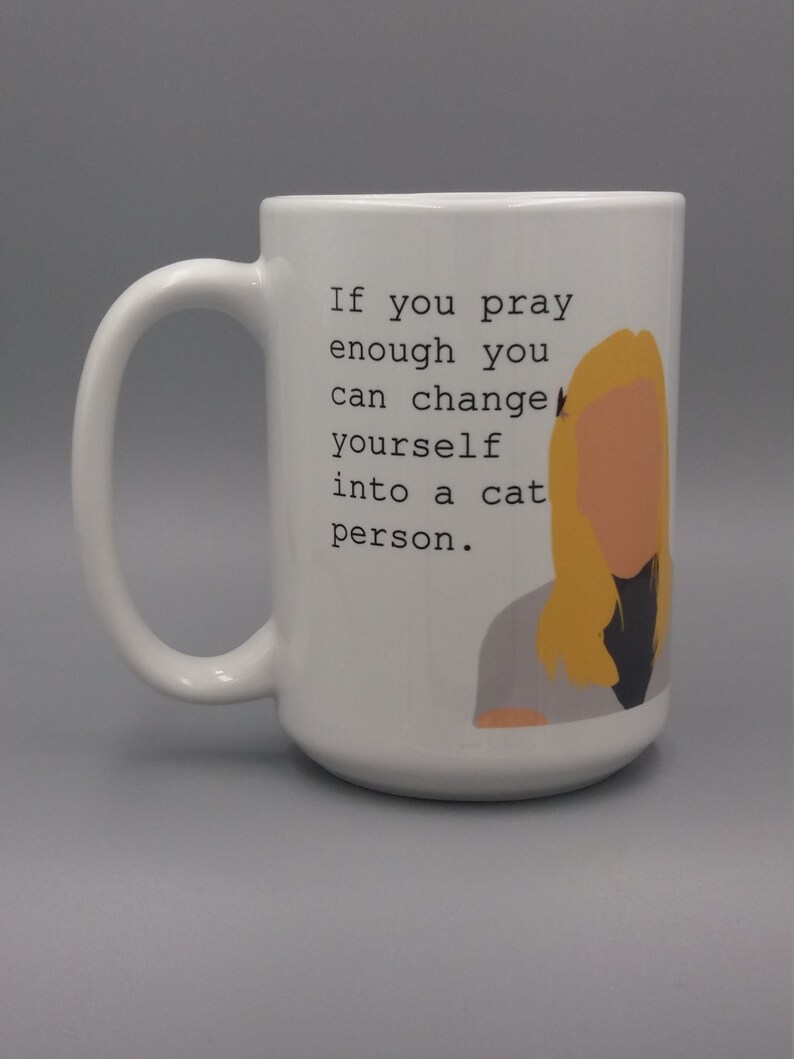 Puede incluir: Taza de cer&aacute;mica blanca con un asa grande. La taza presenta el texto "If you pray enough you can change yourself into a cat person." y una ilustraci&oacute;n de dibujos animados de una persona con cabello rubio.
