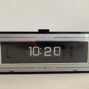 May include: A vintage General Electric digital clock displaying 10:20. The clock has a black rectangular frame with a silver and black face. The words "LIGHTED DIAL" and "GENERAL ELECTRIC" are printed on the face.