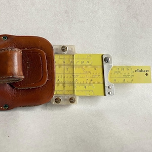 May include: A vintage Pickett slide rule with a yellow scale and clear plastic components. The slide rule is partially housed in a brown leather case with a belt loop. The Pickett logo is visible on the scale.