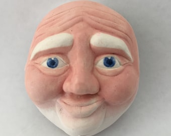 May include: A ceramic face sculpture with a smiling expression. The face is painted with a pinkish skin tone, white eyebrows, and blue eyes. The sculpture has a detailed, aged appearance.