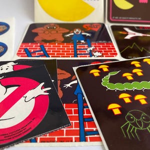 May include: A collection of vintage stickers featuring various designs. Included are stickers with the Ghostbusters logo, E.T., and other cartoon characters. The stickers are square and rectangular, with vibrant colors and detailed illustrations.