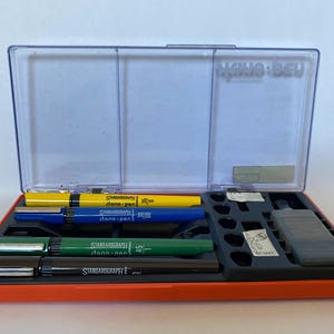 May include: An orange plastic case with a clear lid containing a set of four black, blue, green, and yellow technical drawing pens. The pens are labeled "Standardgraph" and "line-pen".
