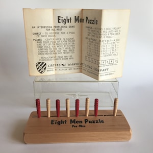 May include: A wooden Eight Men Puzzle game with red and natural wood colored pegs. The game is set up on a wooden board with a label that reads "Eight Men Puzzle Pee-Wee". The game instructions are printed on a folded piece of paper.