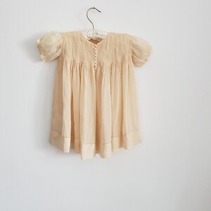 May include: A pale yellow baby dress with puffed sleeves and a button-up front. The dress is hanging on a white hanger against a white wall.