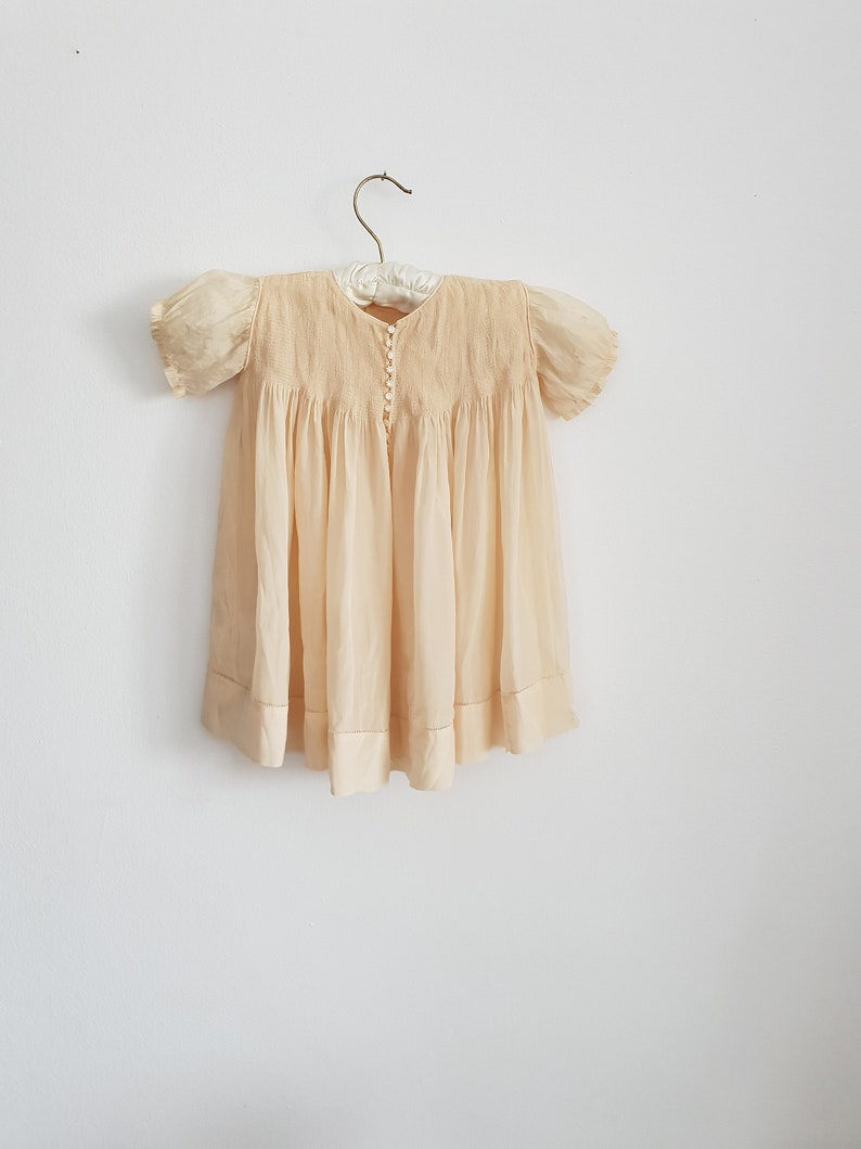 May include: A pale yellow baby dress with puffed sleeves and a button-up front. The dress is hanging on a white hanger against a white wall.