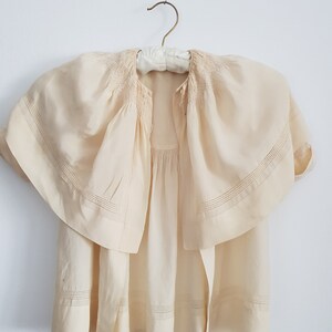 May include: A cream-colored, lightweight, sheer blouse with a gathered neckline and a wide, flowing collar. The blouse has a simple, elegant design with delicate stitching details.