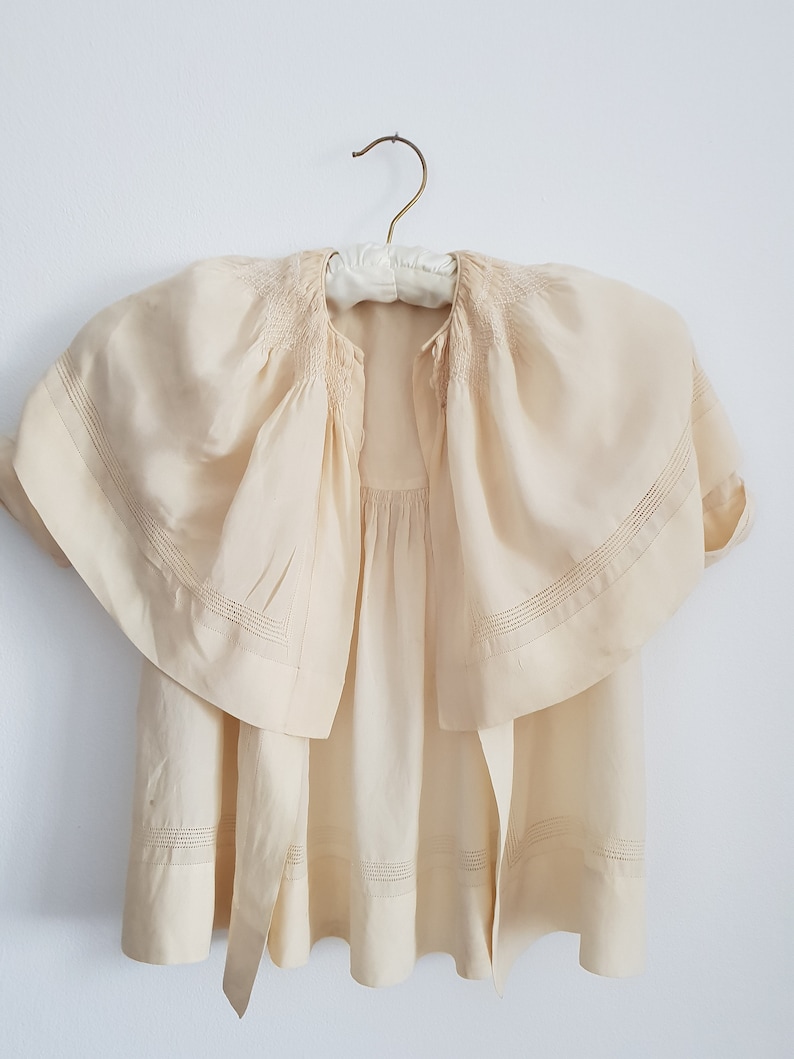 May include: A cream-colored, lightweight, sheer blouse with a gathered neckline and a wide, flowing collar. The blouse has a simple, elegant design with delicate stitching details.