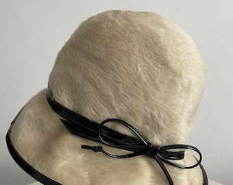 May include: A vintage cloche hat with a cream-colored fur-like texture. The hat features a black patent leather band around the brim and a matching bow detail. The hat is displayed on a red stand.