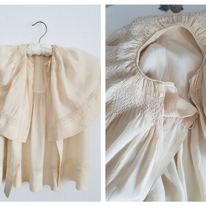 May include: A cream-colored, vintage baby dress with a smocked neckline and a large, ruffled collar. The dress has a tie at the waist.