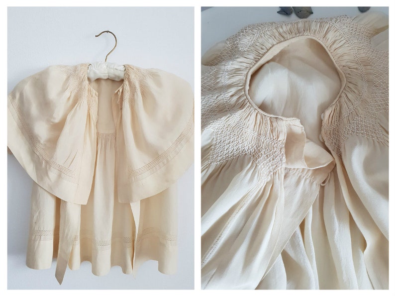 May include: A cream-colored, vintage baby dress with a smocked neckline and a large, ruffled collar. The dress has a tie at the waist.