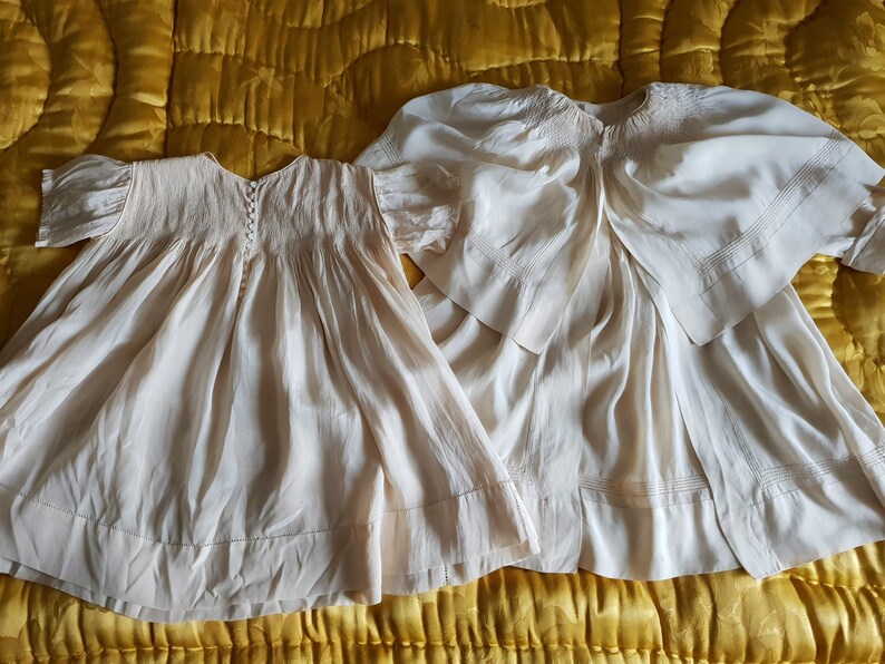 May include: Two vintage white cotton baby dresses with smocking and lace details. The dresses are laid out on a yellow quilted fabric.