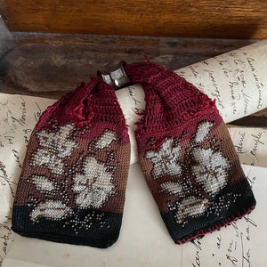 Könnte beinhalten: Ein Paar antike, mit Perlen besetzte und gehäkelte Armbänder mit Blumenmuster. Die Armbänder sind aus rotem, braunem und schwarzem Garn gefertigt und mit weißen Perlen verziert.