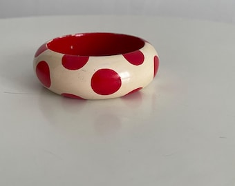 May include: A wooden bangle bracelet with a cream-colored exterior and red polka dots. The interior of the bracelet is red. This bold, round bracelet is a fun accessory.