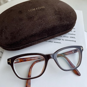 Puede incluir: Gafas de vista de carey marrón con lentes transparentes y detalles dorados. Las gafas están en una caja de terciopelo marrón con las palabras "TOM FORD" grabadas en la parte superior. Las gafas tienen el texto "1-1.6 TOM FORD MADE IN ITALY" en el lateral.
