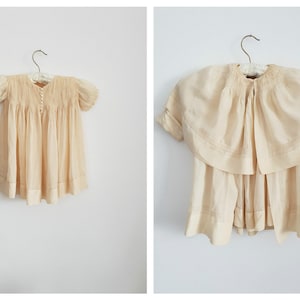 May include: A vintage cream-colored baby dress with a smocked bodice and short sleeves. The dress has a lace trim and is hanging on a wooden hanger.