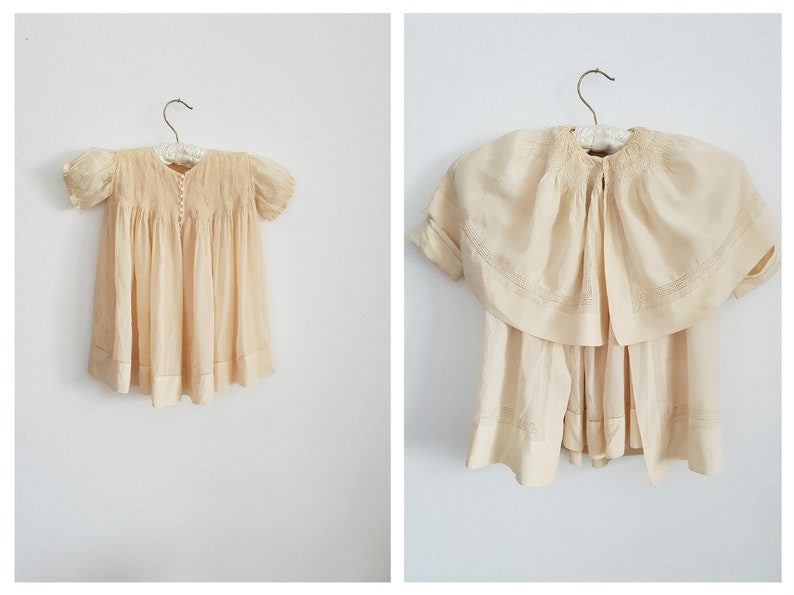 May include: A vintage cream-colored baby dress with a smocked bodice and short sleeves. The dress has a lace trim and is hanging on a wooden hanger.