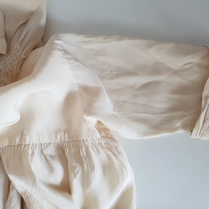 May include: A cream-colored blouse with a gathered neckline and a short sleeve. The sleeve has a stitched trim around the edge.
