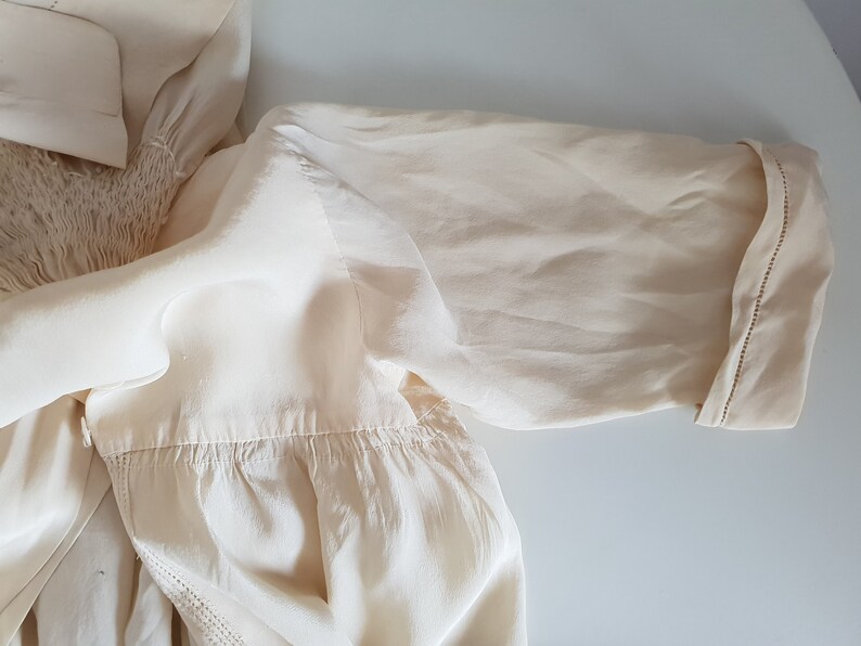 May include: A cream-colored blouse with a gathered neckline and a short sleeve. The sleeve has a stitched trim around the edge.
