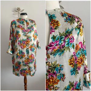 Pode incluir: Vestido vintage com estampa floral, mangas compridas e gola mandarim. O vestido apresenta uma frente abotoada e é adornado com um padrão vibrante de flores rosa, laranja, azul e amarelo sobre um fundo creme. O vestido é exibido em um manequim.