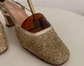Puede incluir: Un par de zapatos de tacón de brillo dorado con una correa de slingback. Los zapatos tienen un tacón de bloque bajo y una punta puntiaguda. El nombre de la marca "Milazzo" es visible en el interior del zapato.
