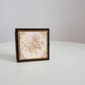 Op de afbeelding: Een klein, vierkant, zwart en gouden compact met een bloemenpatroon aan de voorkant.