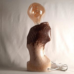 Può includere: Lampada artigianale con base in ceramica marrone e beige e una lampadina ambrata. La base ha una forma organica strutturata. La lampada è collegata con un cavo bianco. La base è alta circa 25 cm.